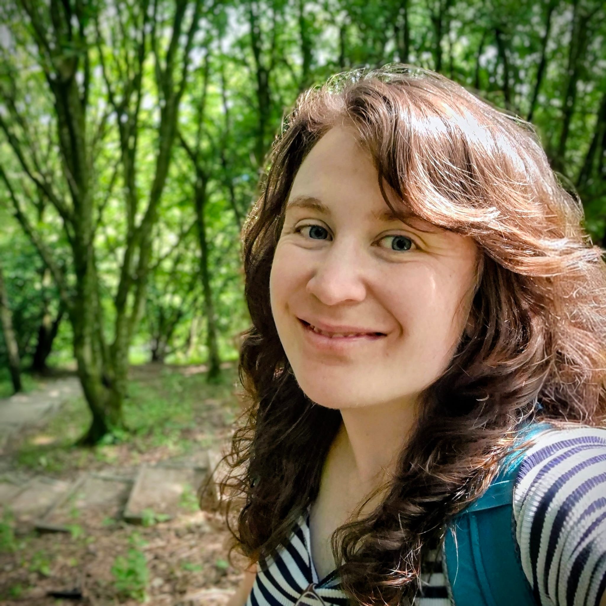 A headshot of Rhian. She is has messy long brown hair and a big smile. She is stood in green woodland.